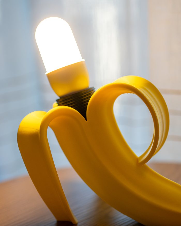 Lampada a forma di banana sbucciata accesa su un tavolo di legno, davanti a una tenda chiara