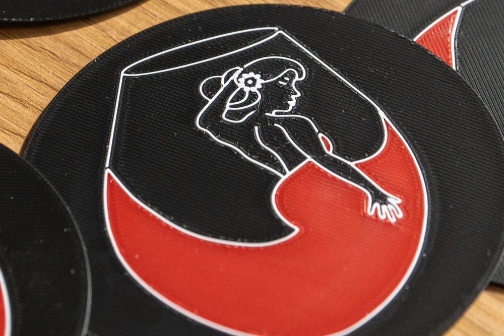 Sottobicchiere in PLA nero visto da vicino, con il disegno di una figura dentro un calice in bianco e rosso