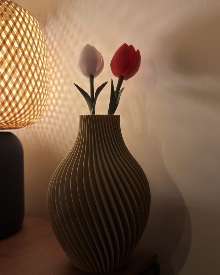 Vaso a coste elicoidali con due tulipani stampati in 3D, uno bianco e uno rosso, accanto a una lampada di vimini accesa